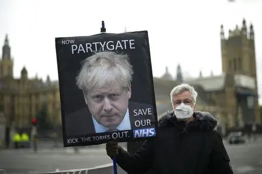 An anti-Conservative Party protester holds a placard with an image of British Prime Minister Boris Johnson including the words "Now Partygate" backdropped by the Houses of Parliament, in London, Wednesday, Dec. 8, 2021. British police are getting ready to issue a first batch of fines on over parties held by Prime Minister Boris Johnson’s staff during coronavirus lockdowns. The Metropolitan Police declined to confirm reports multiple U.K. media outlets that fines would come as soon as Tuesday, 