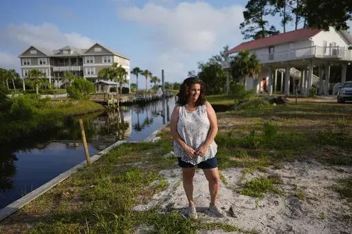 Lisa Bregenzer poses for a picture on the canal-side lot where her home stood until the passage of last year's Hurricane Idalia, Thursday, May 9, 2024, in Horseshoe Beach, Fla. In July, nearly a year after the storm that upended her and her husband's lives, Bregenzer has finally found hope. Her family was approved for a program that will help them build a new home, and if all goes as planned, they'll move into a stilted two-bedroom house on their lot by year's end. (AP Photo/Rebecca Blackwell)
