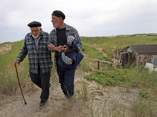 Artist Salvatore Del Deo walked around the shack, right rear, with the aid of his son Romolo Del Deo, on June 27, 2023, in Provincetown, Mass. The 95-year-old painter and his family who were threatened with eviction from their dune shack at the Cape Cod National Seashore in Mass., have won a reprieve. Lawyers for the painter worked out a deal with federal officials allowing Del Deo and his family to continue living in the shack for another five years. (David L Ryan/The Boston Globe via AP)